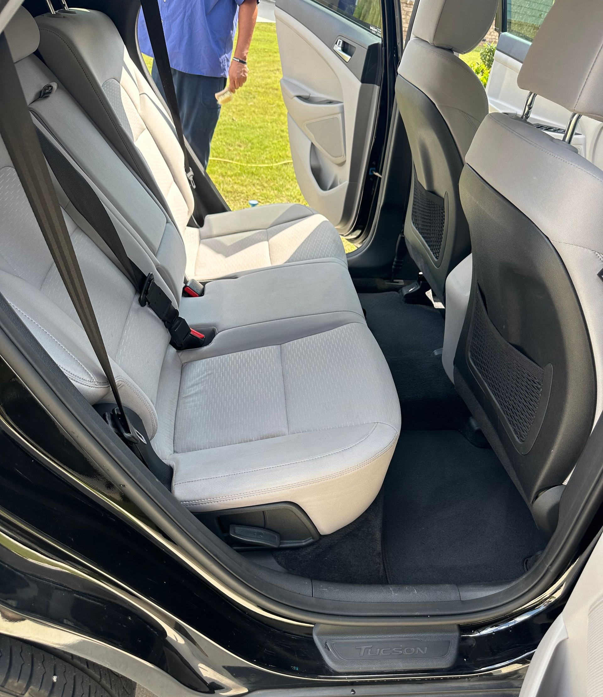 View of the open back seat of a black car with light gray seats and a person partially visible in the background.