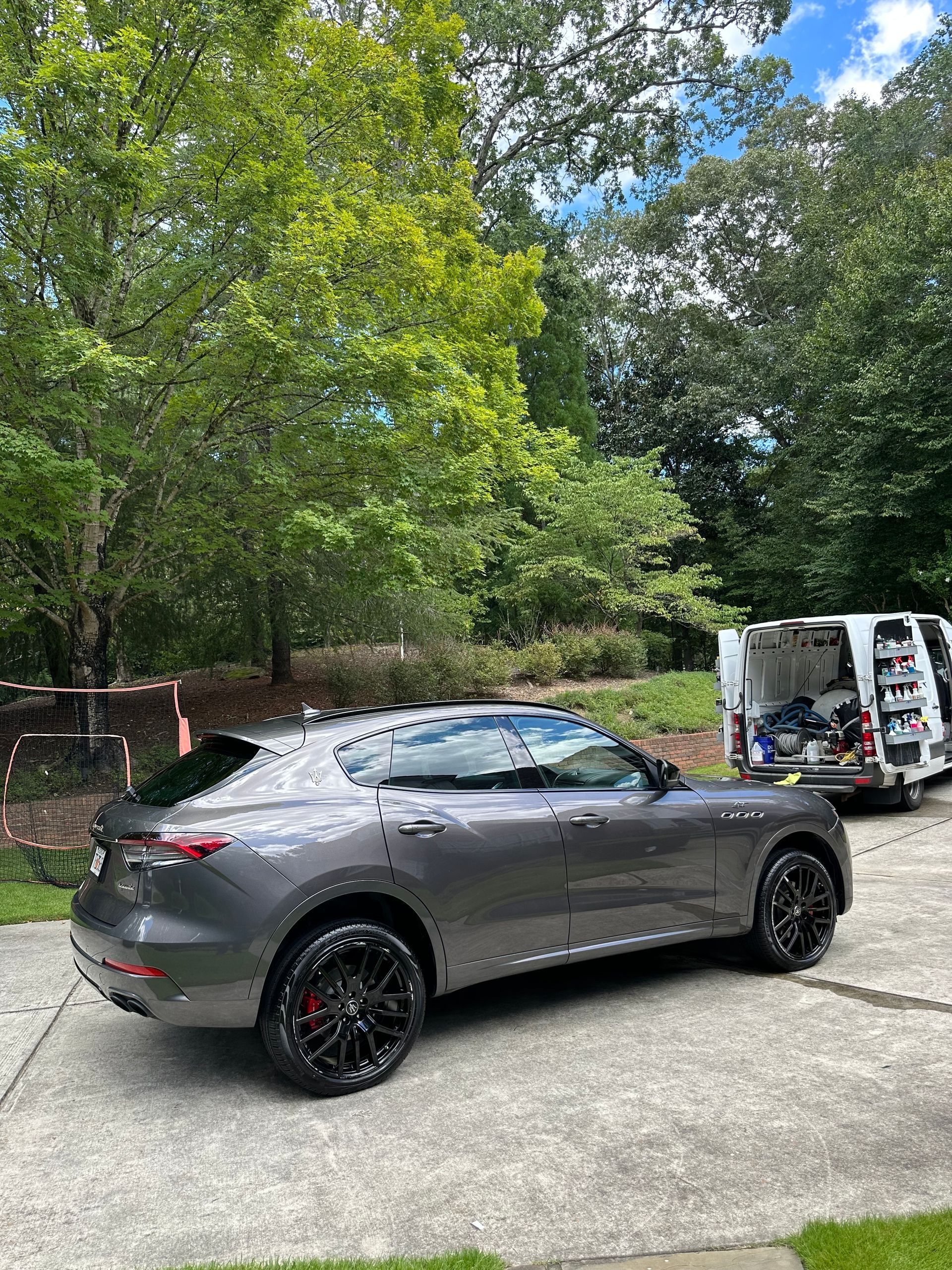 Gray Maserati SUV parked on a concrete driveway with a van in the background, surrounded by trees.