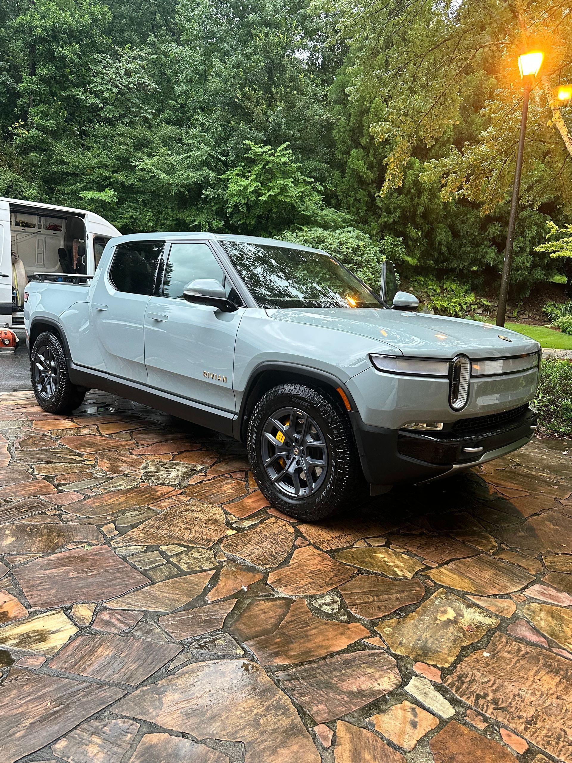 Light blue Rivian electric pickup truck parked on a wet stone driveway, forest background.