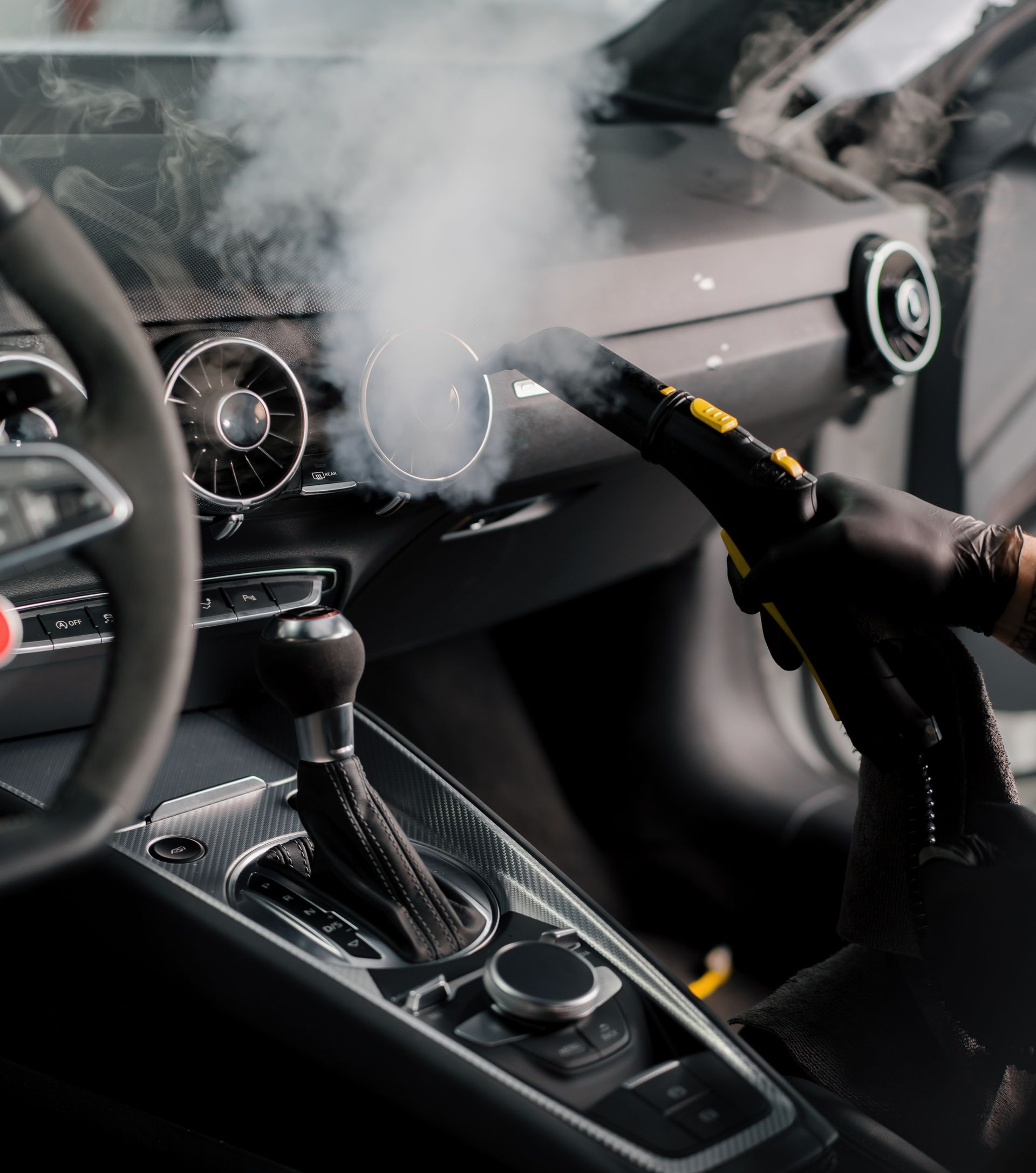 Car interior being steam cleaned, focus on the dashboard and vents, with steam visible.
