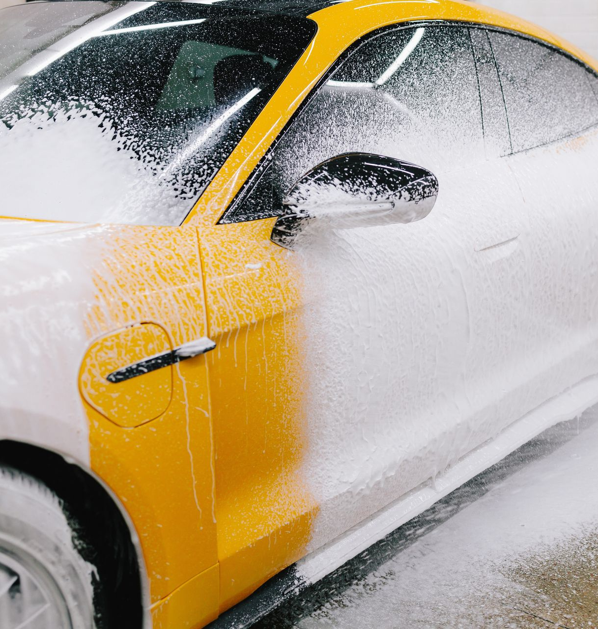 Yellow and white car being washed with thick white foam, in a garage.