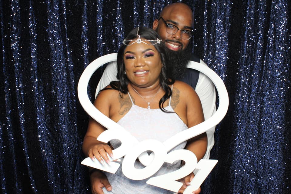 Two people posing before a dark sequin backdrop, holding a white decorative prop and smiling.