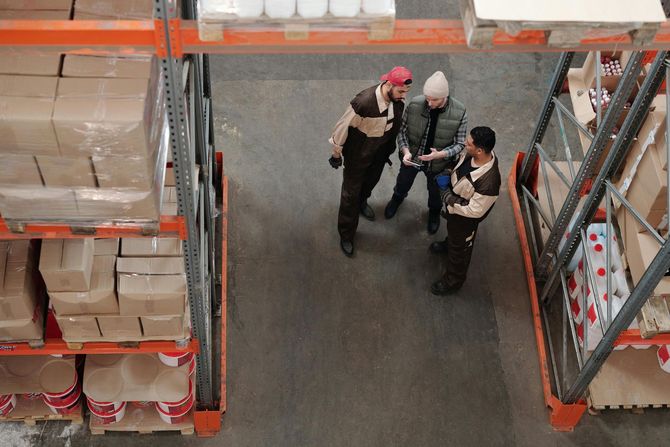 Warehouse workers reviewing inventory, standing between shelves of packaged goods.