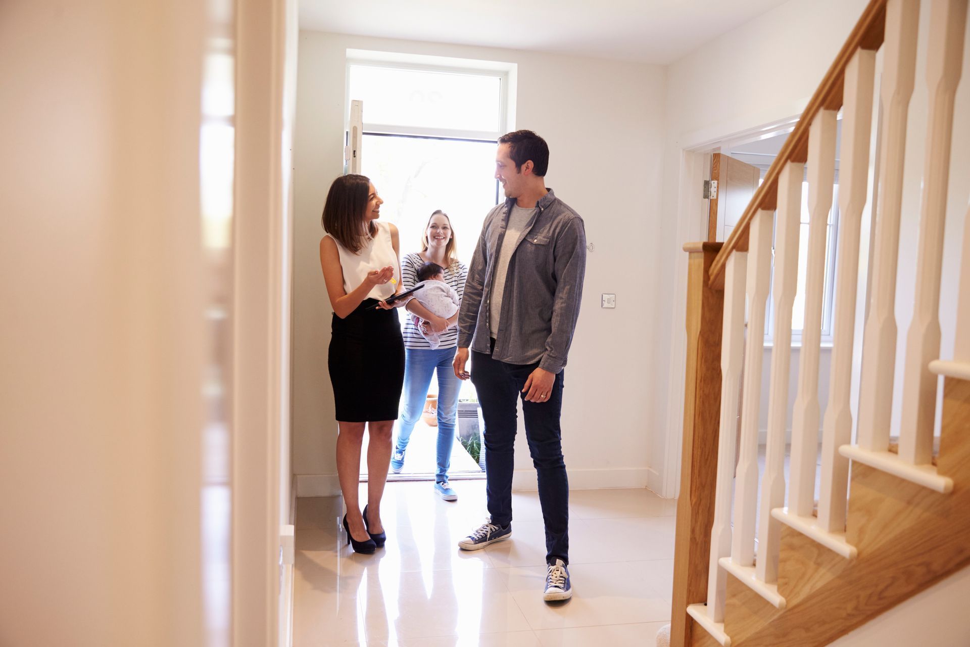 Real estate agent showing a couple a home; bright entry with stairs and open front door.