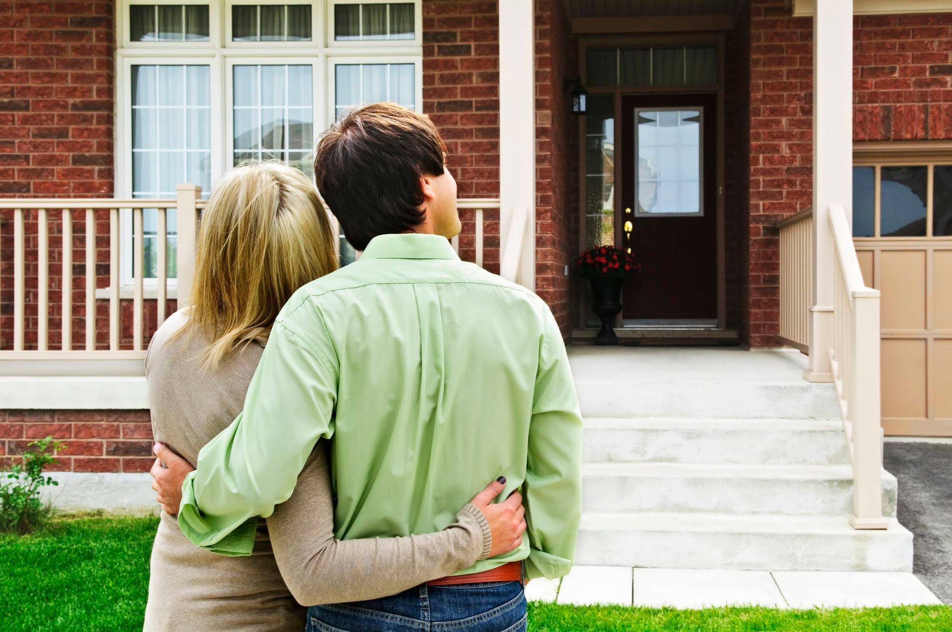 Couple hugging in front of a brick house with white trim; looking towards the front door.