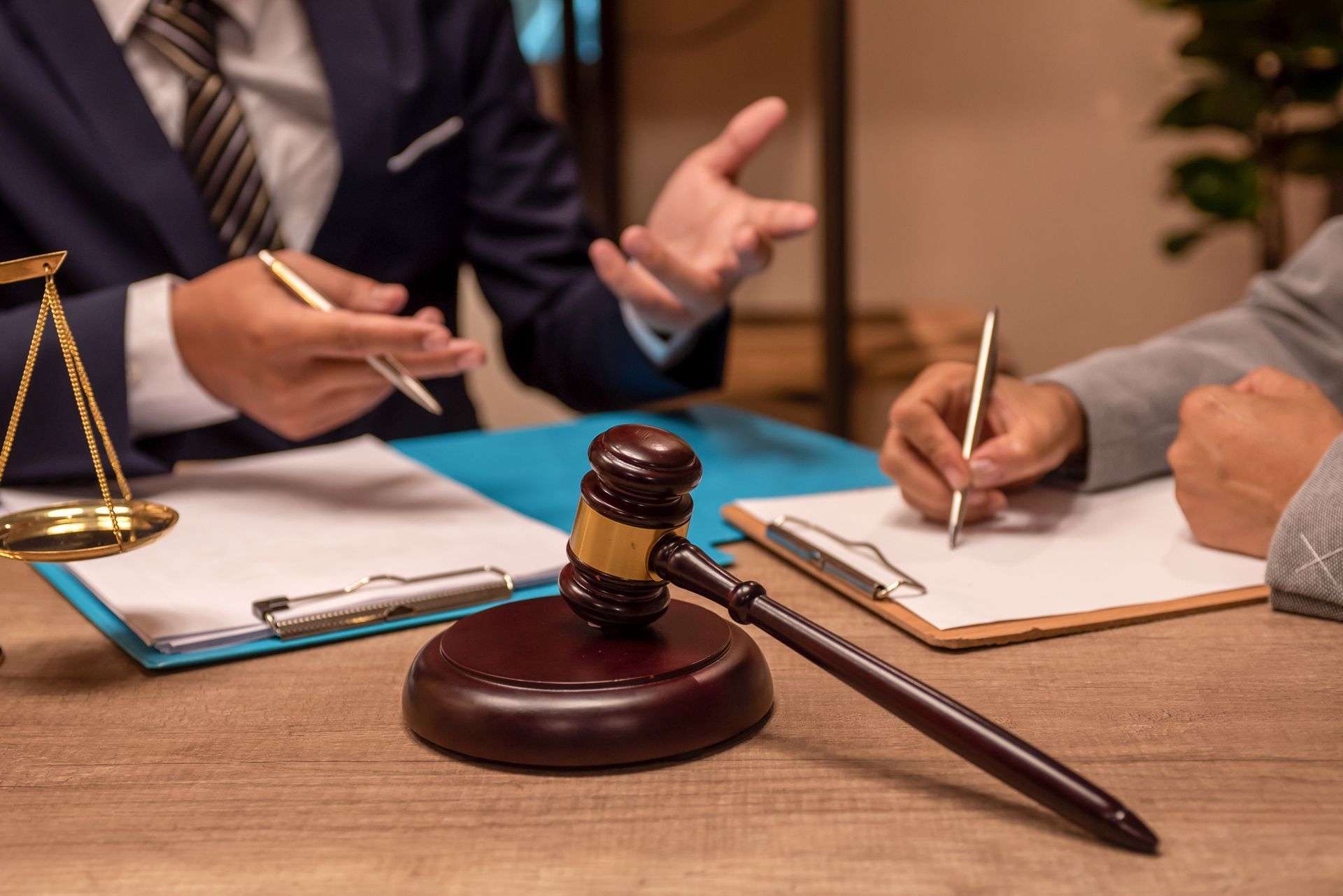 Gavel on table with two people in suits at a desk discussing documents. Scales of justice in background.