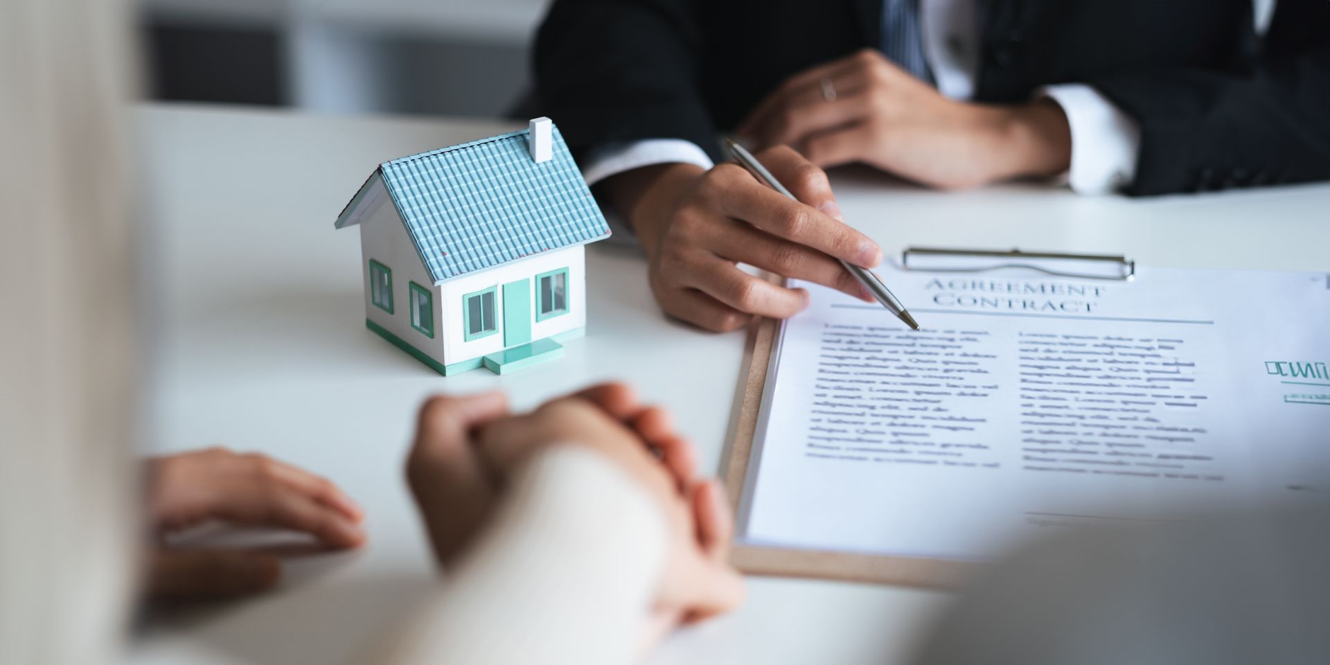 A person in a suit points at a contract. A small house model sits on the table. Someone's hands are clasped.