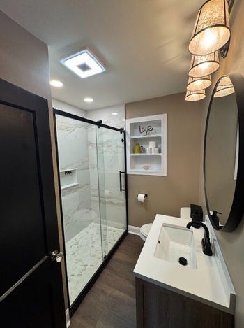 Modern bathroom with glass shower, dark flooring, white sink vanity, and beige walls illuminated by pendant lights