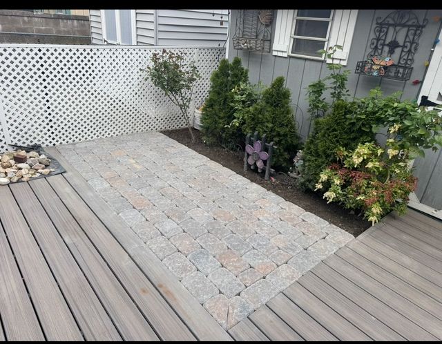 Backyard deck with gray pavers, wood planks, white lattice fence, and potted shrubs along the edge