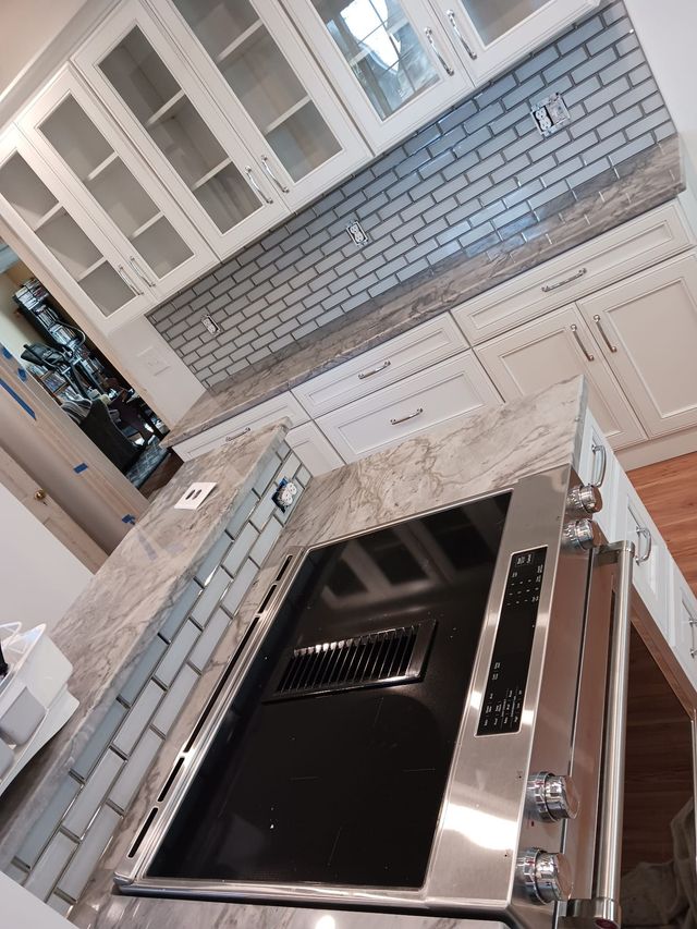 Kitchen with a black stovetop and stainless steel hood beneath white cabinets and a light blue tile backsplash