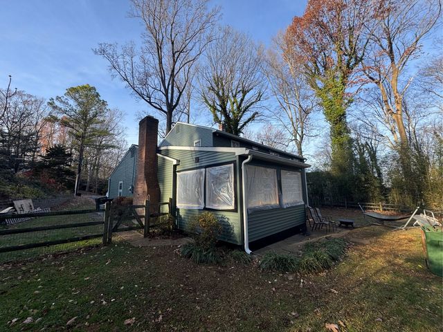 Small green house with chimney in a leafy yard, surrounded by bare trees in autumn light