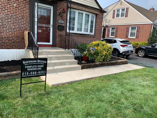 Brick house front entrance with steps, sign, shrubbery, and parked cars in a suburban yard.
