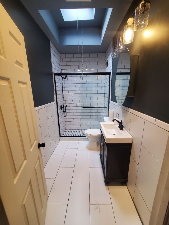 Long narrow bathroom with white tile, glass shower at back, vanity sink, and dark accent wall ceiling.