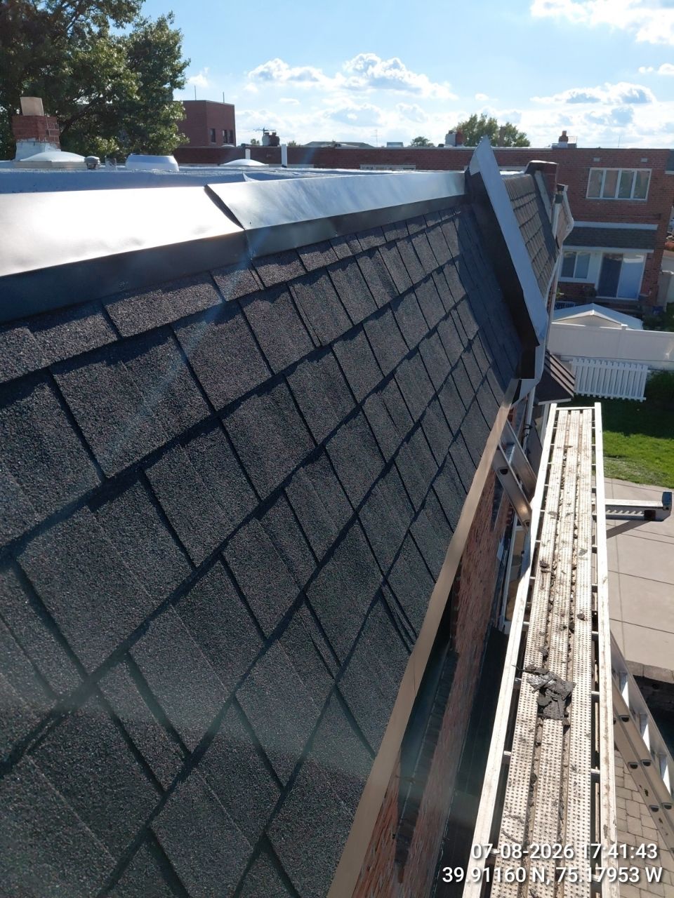 Steep shingled roof under repair with scaffolding, blue sky, and nearby brick buildings