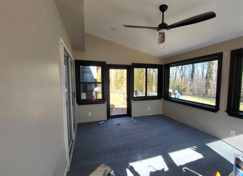 Empty sunroom with gray carpet, black ceiling fan, and large windows letting in bright sunlight.