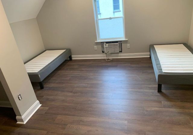 Empty bedroom with two single beds, brown wood floor, and a small window air conditioner beneath the window