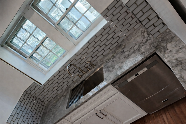Tilted kitchen interior with white cabinets, dark countertop, gray tile backsplash, and bright windows