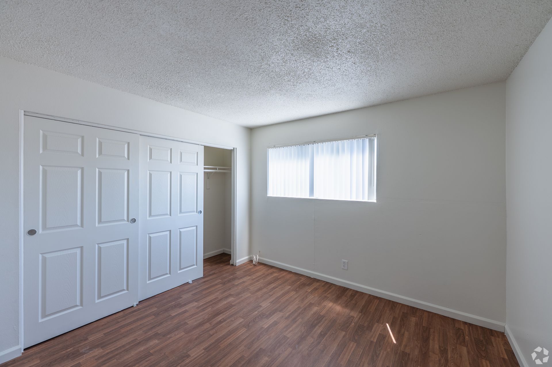 Empty bedroom with white walls, wood-look floor, closet doors, and a window with vertical blinds
