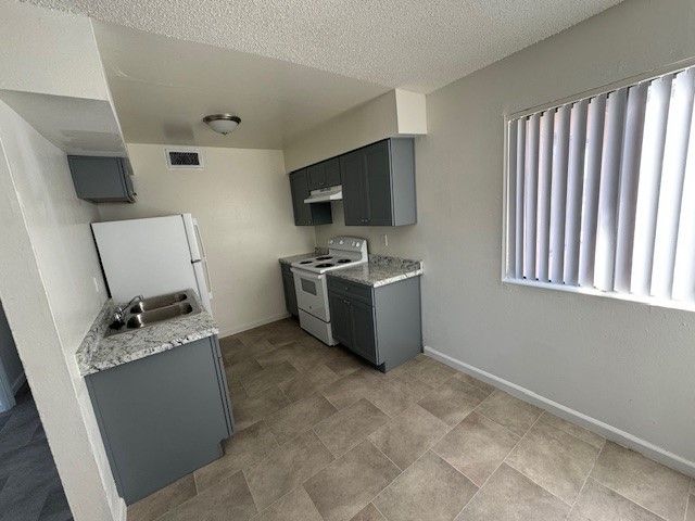Empty apartment kitchen with gray cabinets, white appliances, and vertical blinds by a window