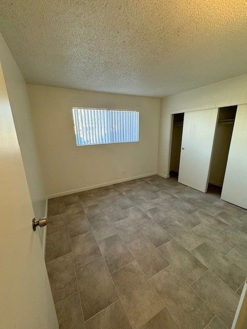 Empty beige bedroom with tiled floor, window blinds, and open closet doors