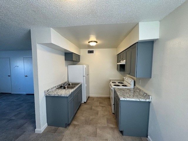 Empty galley kitchen with blue cabinets, granite counters, white appliances, and tiled floor.