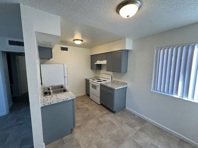 Small empty kitchen with gray cabinets, white appliances, granite counters, and vertical blinds by the window