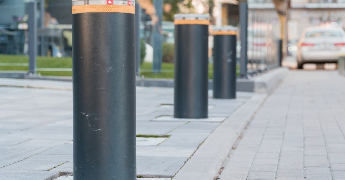 A row of dark gray cylindrical security bollards with an orange stripe on top installed along a brick sidewalk.