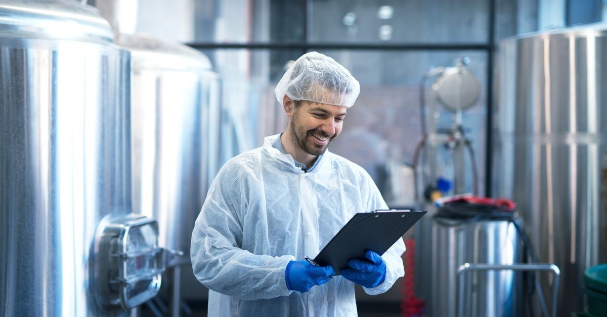 A smiling industrial worker in protective gear checks a clipboard.