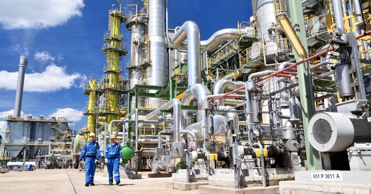 Two men in hard hats and jumpsuits walk alongside a large chemical plant. Steel pipes and machinery 