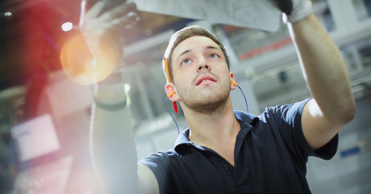 A man holds a thin piece of steel up to the light. He has a pencil tucked behind one ear and is wearing headphones.