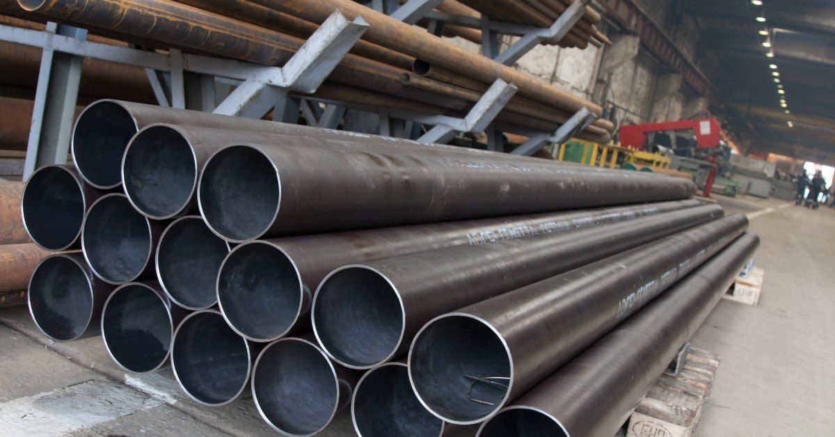 Three rows of steel pipes are stacked on top of wooden blocks on a concrete floor. Rusty pipes are stored nearby.