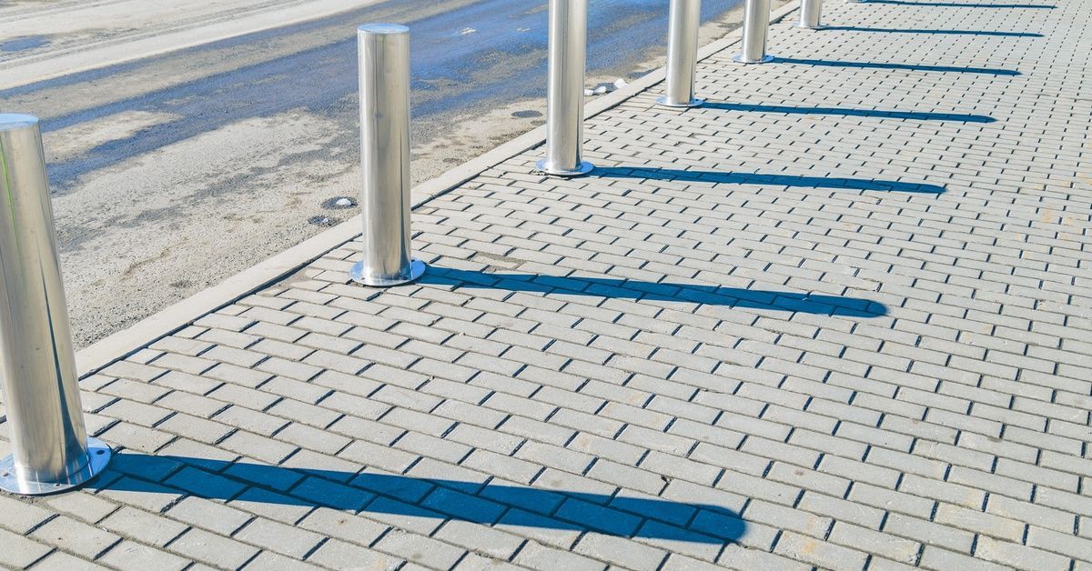 A row of stainless steel bollards lining an empty cobblestone sidewalk. Each bollard casts a long shadow.