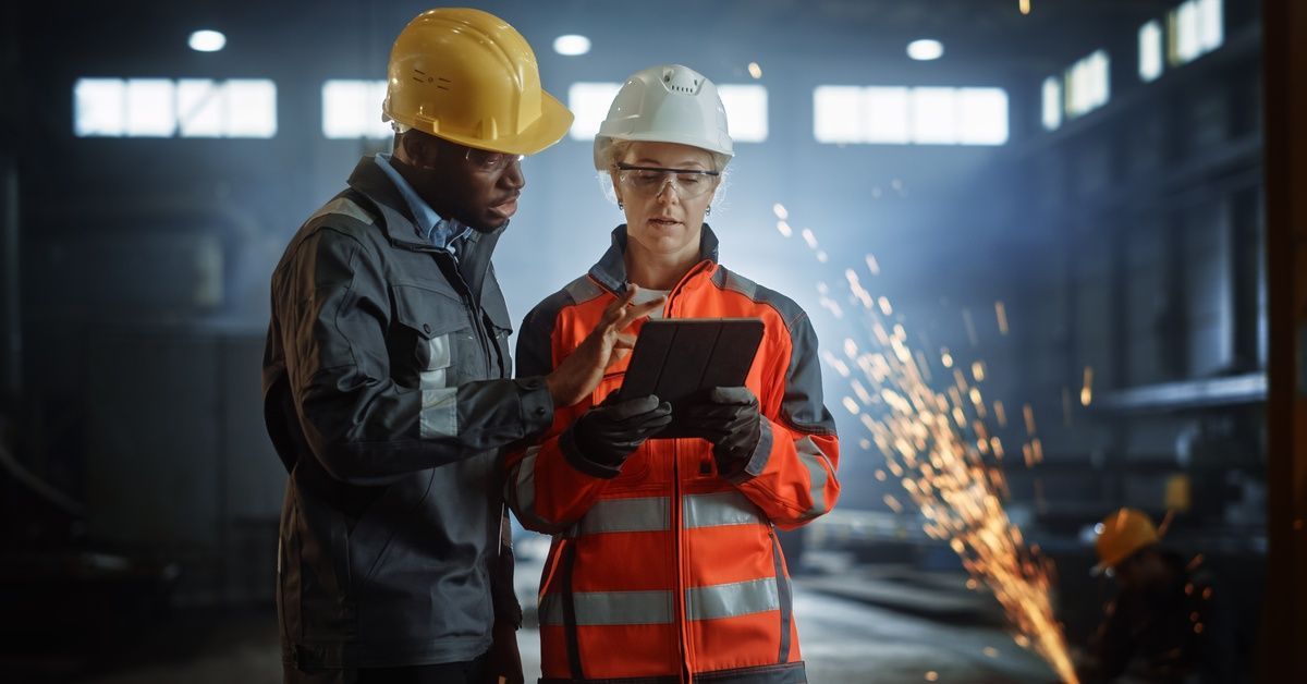 Two workers in hard hats examining a tablet while a welder in the background sends orange sparks int
