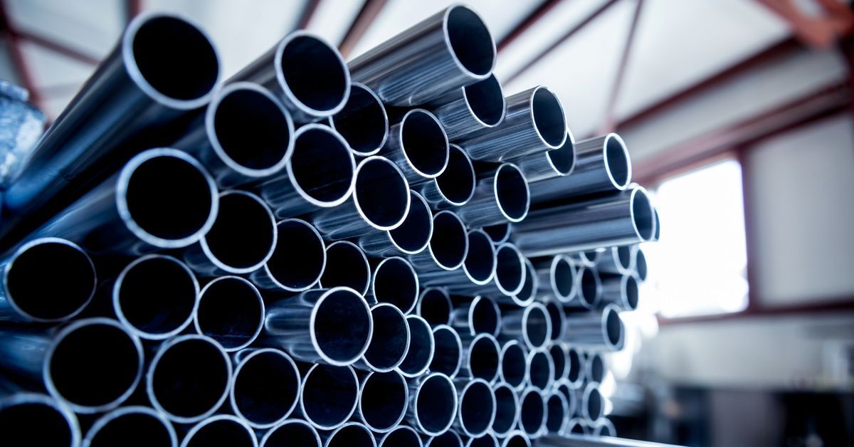 Stainless steel pipes stacked in rows inside a room with red beams on the ceiling. Sunlight enters through a window.