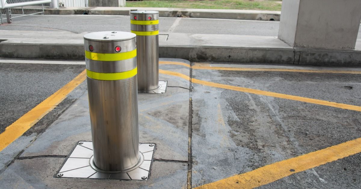 Two stainless steel bollards with surface mounting plates stand on concrete near a roadway.