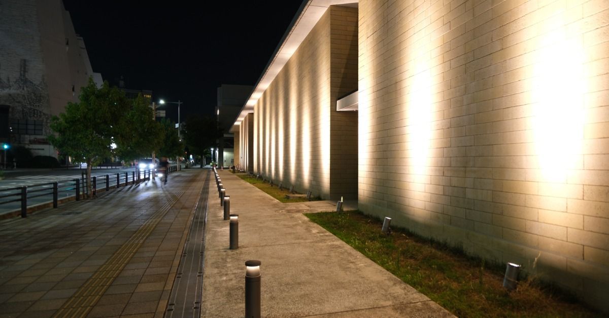 Modern commercial building exterior illuminated at night, with a wide sidewalk and streetlights lini