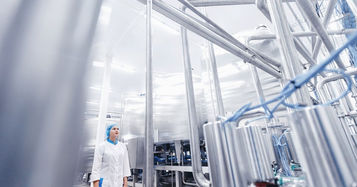 A worker in protective clothing stands among stainless steel tanks and pipes inside a clean industri