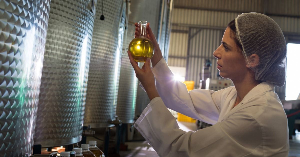 A technician inspects a liquid sample beside large dimpled stainless steel tanks inside a processing facility.