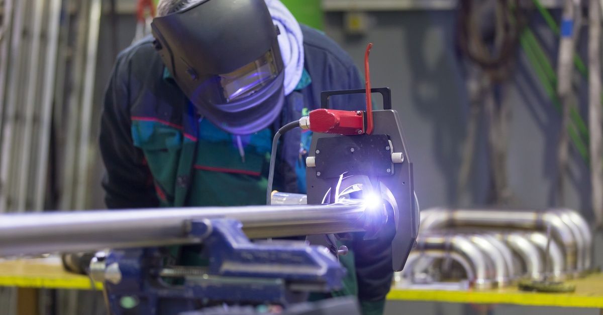 An industrial operator wearing protective gear observes an orbital welding machine inside a metal manufacturing shop.