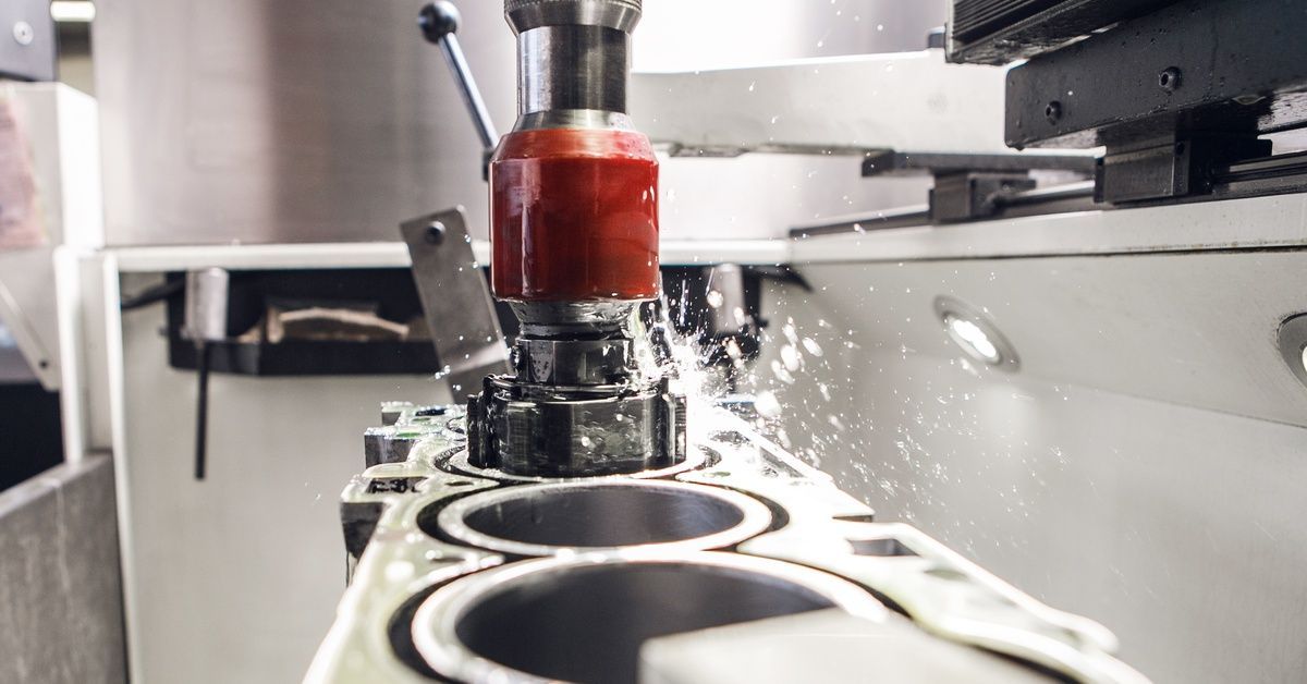 A precision milling machine drills into a metal engine block, spraying coolant and fine metallic shavings.