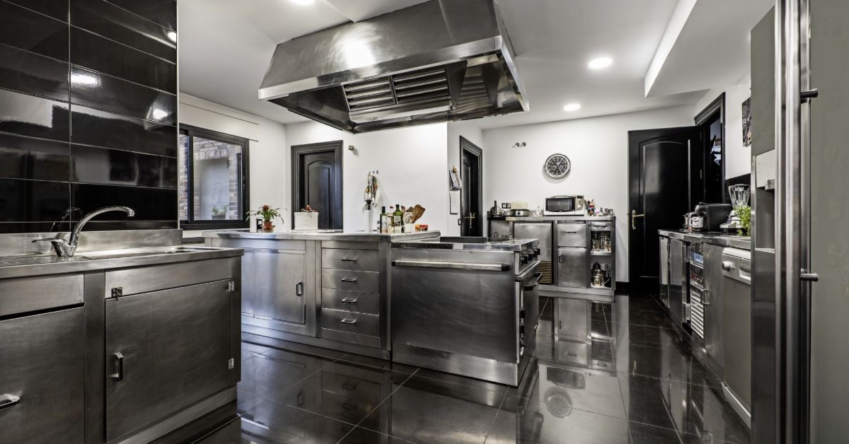 Modern residential kitchen with stainless steel cabinets, a large range hood, a central island, and black tile flooring.