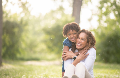Woman smiles, hugging a child outdoors in a park. Both are laughing.