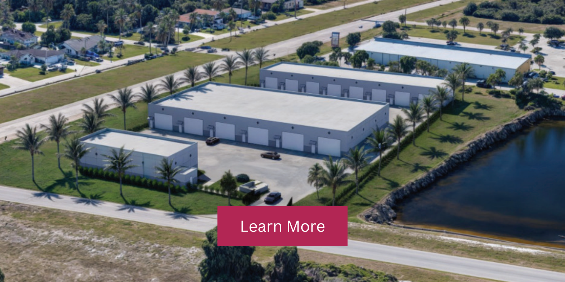 Aerial view of a white industrial building complex beside a canal, with palm trees and a Learn More button.