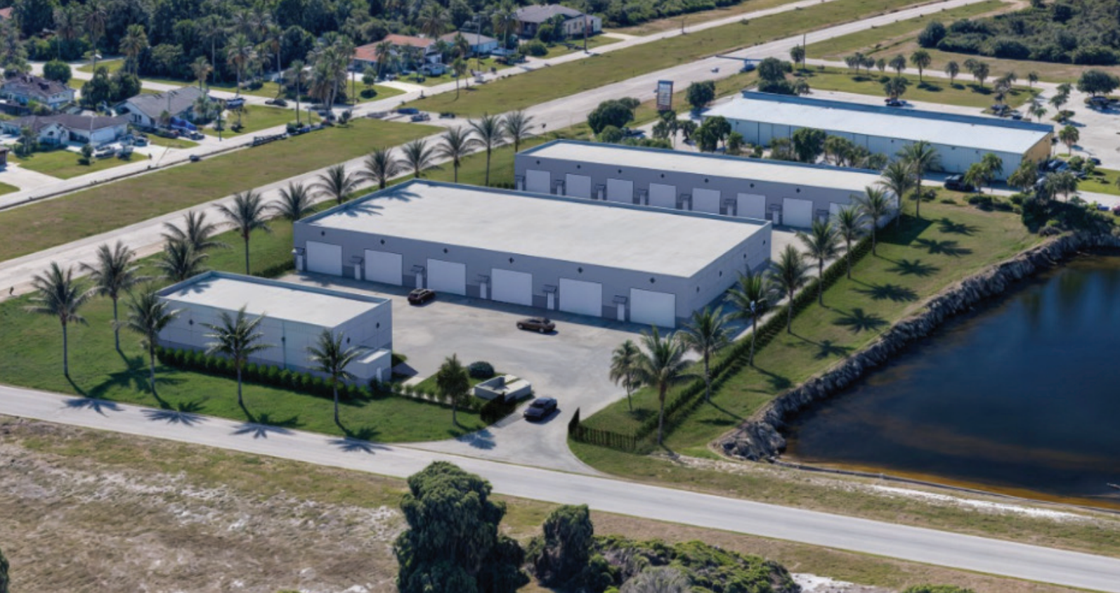 Aerial view of a warehouse complex with parking lots beside a pond and road.