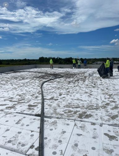 Workers in neon vests on a white rooftop under a cloudy blue sky, with a hose across the surface