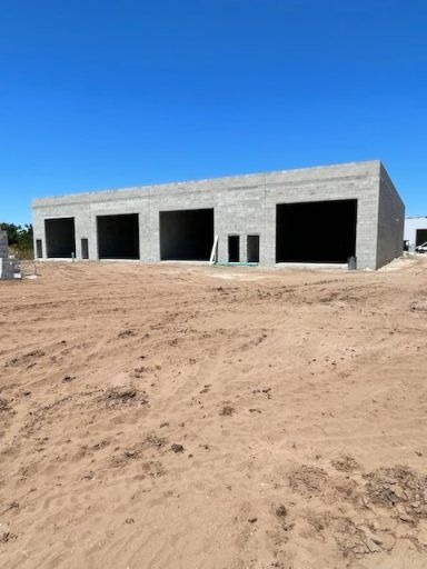 Unfinished gray concrete building with open garage bays on a dirt lot under a clear blue sky