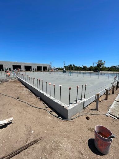Concrete foundation slab under construction at a site, with rebar rods around the edges and a building in the background
