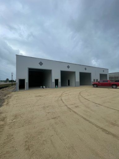 White industrial building with three open bays on a sandy lot under a cloudy sky, red truck parked at right
