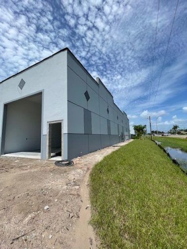 Industrial warehouse exterior with white walls, open garage door, gravel lot, and grassy roadside under a cloudy sky