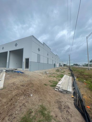 Construction site beside a white industrial building under cloudy skies, with dirt, materials, and fencing in front.